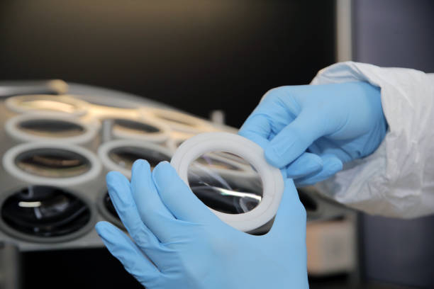 Gloved hands holding a clear circular optical element above a tray